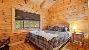A cozy wooden cabin bedroom with a bed, a window with blinds, a bedside table with a lamp, and a chair.