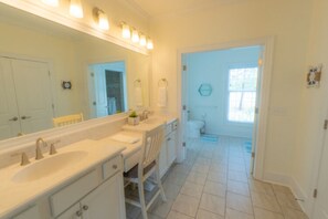 Une salle de bain avec un double vasque, un grand miroir et un meuble-lavabo avec des tiroirs. Une porte donne accès à l’espace douche.