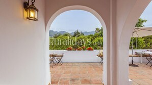 An outdoor patio with a view of mountains, a white wall with a lantern, and a seating area with a table and chairs.