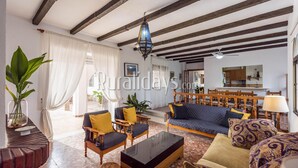 A spacious living room with a sofa, armchairs, and a coffee table. There is a dining area with wooden furniture and a chandelier.
