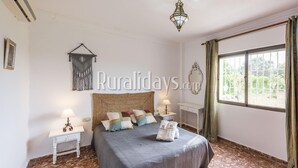 A bedroom with a large bed, two bedside tables, a hanging macrame decoration, a chandelier, and a window with curtains.