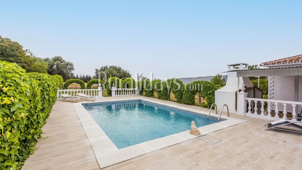 A rectangular swimming pool with a white deck, surrounded by a white fence and lush greenery.