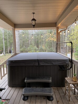 A hot tub on a wooden deck with a view of trees, a step stool, and string lights.