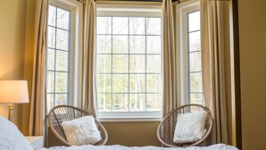 A bedroom with a large window, two wicker chairs, a bed with a white bedspread, and a nightstand with a lamp. - Big Beautiful Georgian Bay Family Cottage (Tiny)