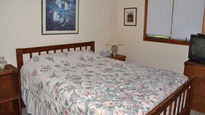 Une chambre avec un lit en bois, une literie à motifs floraux, une table de chevet avec une lampe, un téléviseur posé sur un meuble en bois et une fenêtre avec des stores.