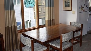 Un coin repas avec une table en bois et des chaises à motifs, une fenêtre avec des rideaux et un réfrigérateur à l’arrière-plan.