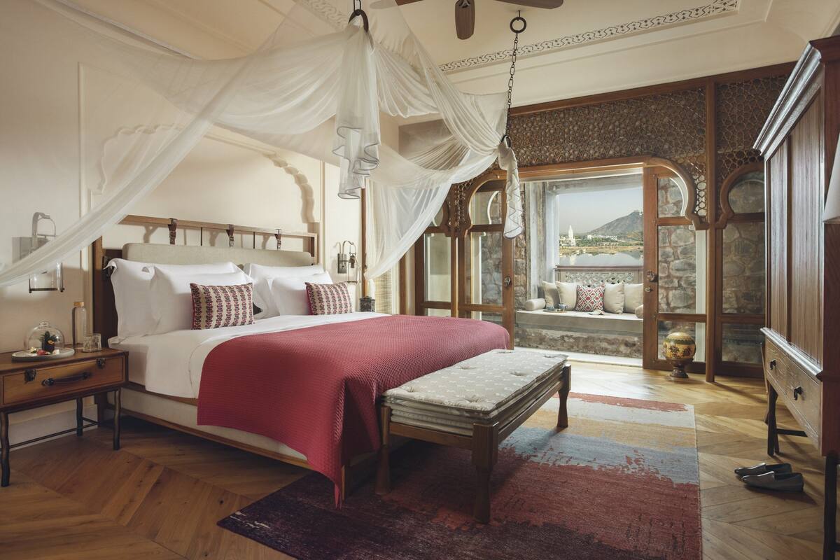 A bedroom with a large bed, a nightstand, a bench, and a view of a mountain through a window.