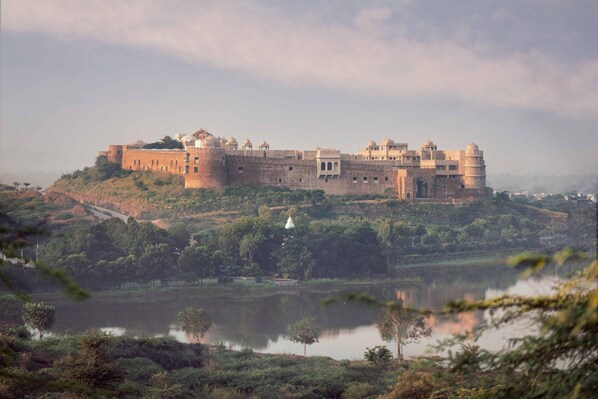 A large, historic castle with multiple towers and a moat, surrounded by lush greenery and a calm lake. - Six Senses Fort Barwara (Chauth Ka Barwara)