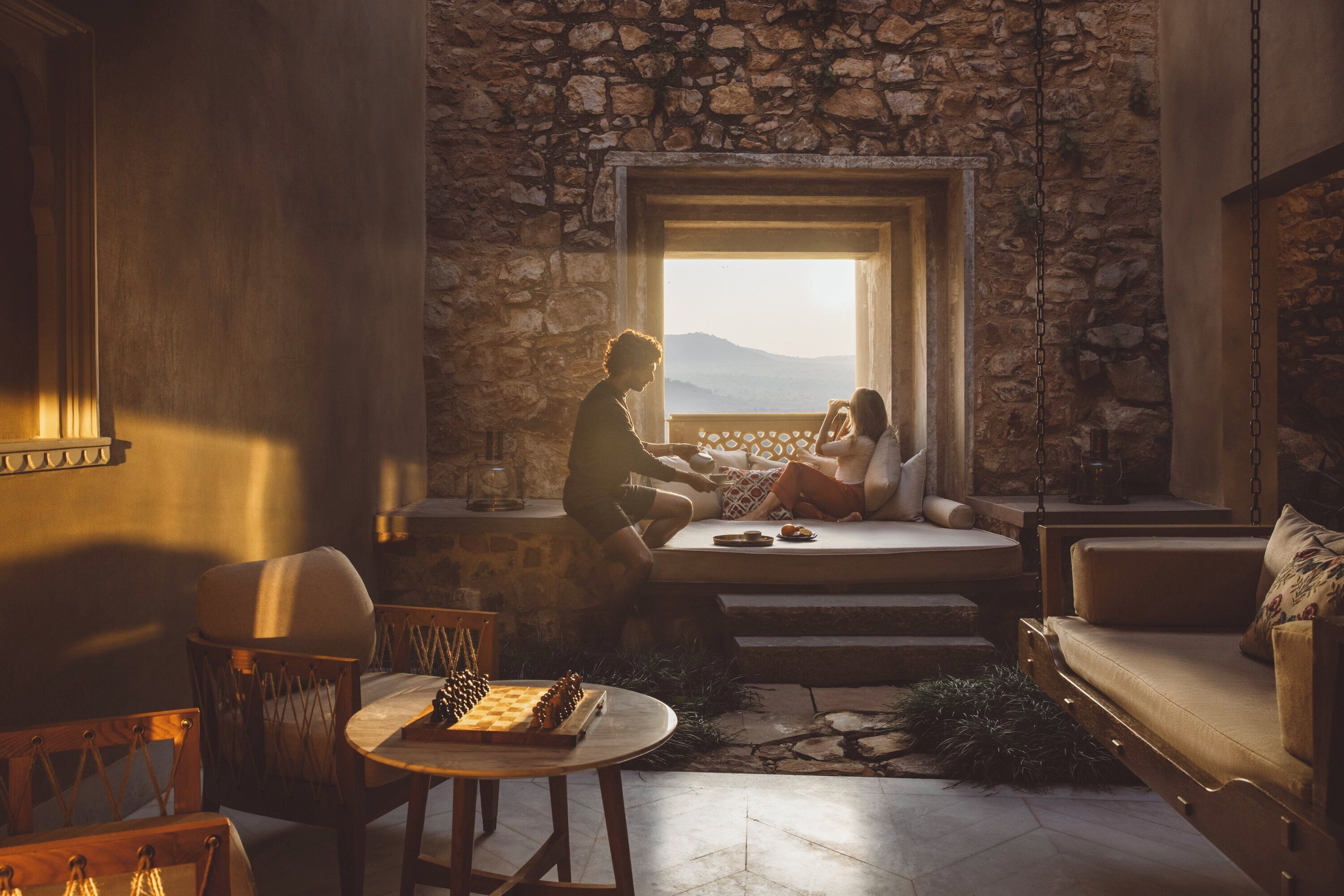 A cozy room with a stone wall, a large window, a chessboard, and a sofa.