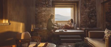 A cozy room with a stone wall, a large window, a chessboard, and a sofa.