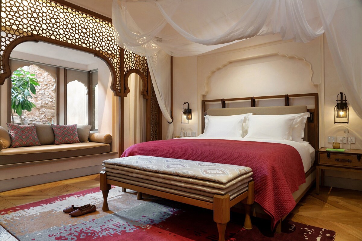 Un dormitorio con una cama grande, un banco, un asiento junto a la ventana y un adornado en la pared.