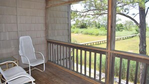 A covered porch with a wooden railing, two white chairs, and a view of a grassy area with trees. - Fowling View | Townhouse w/ Pier, Crabbing Dock & Sunset Views (Chincoteague)