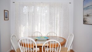 A dining area with a round table and four white chairs, a wooden floor, and a window with sheer curtains. - Fowling View | Townhouse w/ Pier, Crabbing Dock & Sunset Views (Chincoteague)