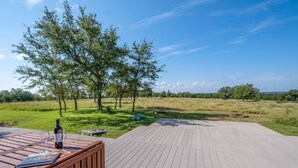 Une terrasse en bois avec une bouteille et des verres, entourée d’une pelouse et de quelques arbres.