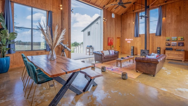 A spacious living room with a wooden ceiling, large windows, a dining table, and a cozy seating area. - Hill Country Barn - Sauna & Cedar Hot Tub (Johnson City)