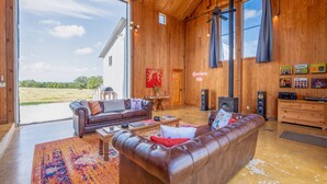 A spacious living room with a large brown leather sofa, a coffee table, and a wooden cabinet. A large window offers a view of a green landscape. - Hill Country Barn - Sauna & Cedar Hot Tub (Johnson City)