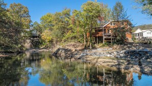 A cabin by a lake with a stone wall and wooden deck, surrounded by trees.