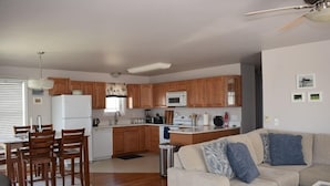Una cocina moderna con armarios de madera, un refrigerador blanco y un microondas. Hay un comedor con una mesa y sillas, y una sala de estar con un sofá beige y almohadones decorativos.