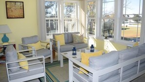 Un salón luminoso con sofá, sillones y una mesa de centro. Amplias ventanas con marcos blancos ofrecen vistas al exterior.