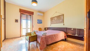 A bedroom with a bed, a chair, a desk, a lamp, and a painting on the wall. - GuestHost - Roomy and bright apartment of 150 sqm, with two balconies and a panoramic terrace of 150 sqm with views of Syracuse, Ortigia and the sea. The flat can accommodate up to 5 people and is located on the 5th floor of a building with lift, in a re (Siracusa)