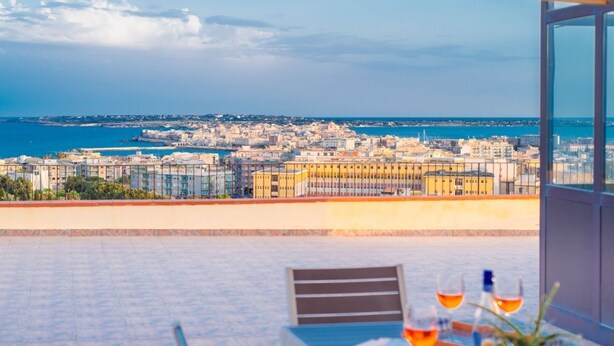 A terrace with a table set for two, overlooking a coastal cityscape. - GuestHost - Roomy and bright apartment of 150 sqm, with two balconies and a panoramic terrace of 150 sqm with views of Syracuse, Ortigia and the sea. The flat can accommodate up to 5 people and is located on the 5th floor of a building with lift, in a re (Siracusa)