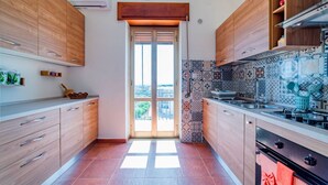 A modern kitchen with wooden cabinets, a tiled backsplash, and a view through a glass door. - GuestHost - Roomy and bright apartment of 150 sqm, with two balconies and a panoramic terrace of 150 sqm with views of Syracuse, Ortigia and the sea. The flat can accommodate up to 5 people and is located on the 5th floor of a building with lift, in a re (Siracusa)