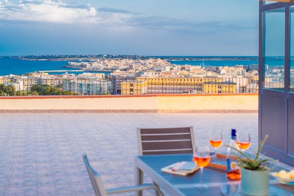 A terrace with a table set for two, overlooking a coastal cityscape.