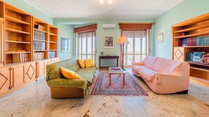 A living room with a sofa, a coffee table, and bookshelves. - GuestHost - Roomy and bright apartment of 150 sqm, with two balconies and a panoramic terrace of 150 sqm with views of Syracuse, Ortigia and the sea. The flat can accommodate up to 5 people and is located on the 5th floor of a building with lift, in a re (Siracusa)