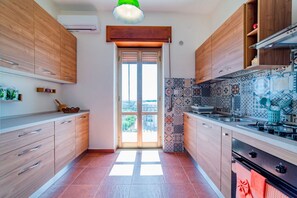 A modern kitchen with wooden cabinets, a tiled backsplash, and a view through a glass door. - GuestHost - Roomy and bright apartment of 150 sqm, with two balconies and a panoramic terrace of 150 sqm with views of Syracuse, Ortigia and the sea. The flat can accommodate up to 5 people and is located on the 5th floor of a building with lift, in a re (Siracusa)
