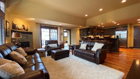 A spacious living room with a large brown leather sofa, a matching ottoman, and a fireplace. There is a kitchen area with dark wooden cabinets and a refrigerator.