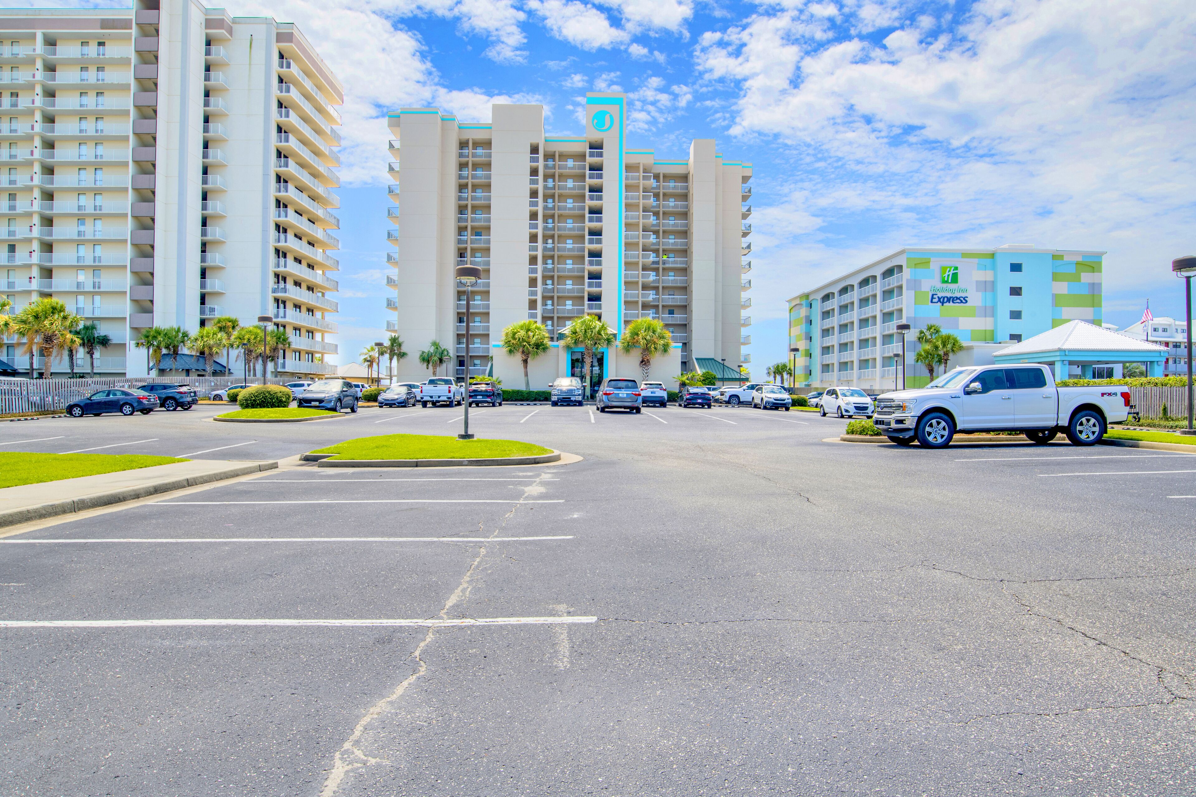 Un hôtel composé de plusieurs immeubles de grande hauteur, un parking rempli de voitures et des palmiers.