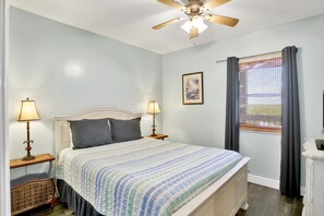 A bedroom with a bed, two bedside lamps, a ceiling fan, a window with curtains, and a framed picture on the wall.