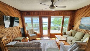L’intérieur d’une cabane est agrémenté de boiseries, d’un ventilateur de plafond, d’un téléviseur fixé au mur, d’un canapé, d’un fauteuil, d’une petite table et offre une vue à travers les portes vitrées.