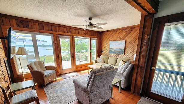 Une pièce dotée d’un plancher en bois, d’un ventilateur de plafond, d’un canapé, d’un fauteuil, d’une table et offrant une vue sur un lac grâce à de grandes fenêtres.