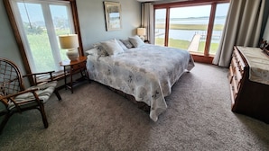 Une chambre à coucher comprenant un lit, une chaise, une commode, une lampe et une fenêtre donnant sur un plan d’eau.