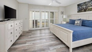 A bedroom with a bed, a television, a dresser, a ceiling fan, and a balcony with shutters.