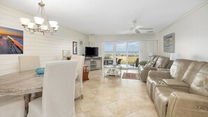 A spacious living room with a dining area, a television, and a large window offering a view of the beach.