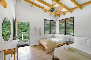 Una habitación con dos camas, una mesa de madera, un ventilador de techo y ventanas grandes con persianas.