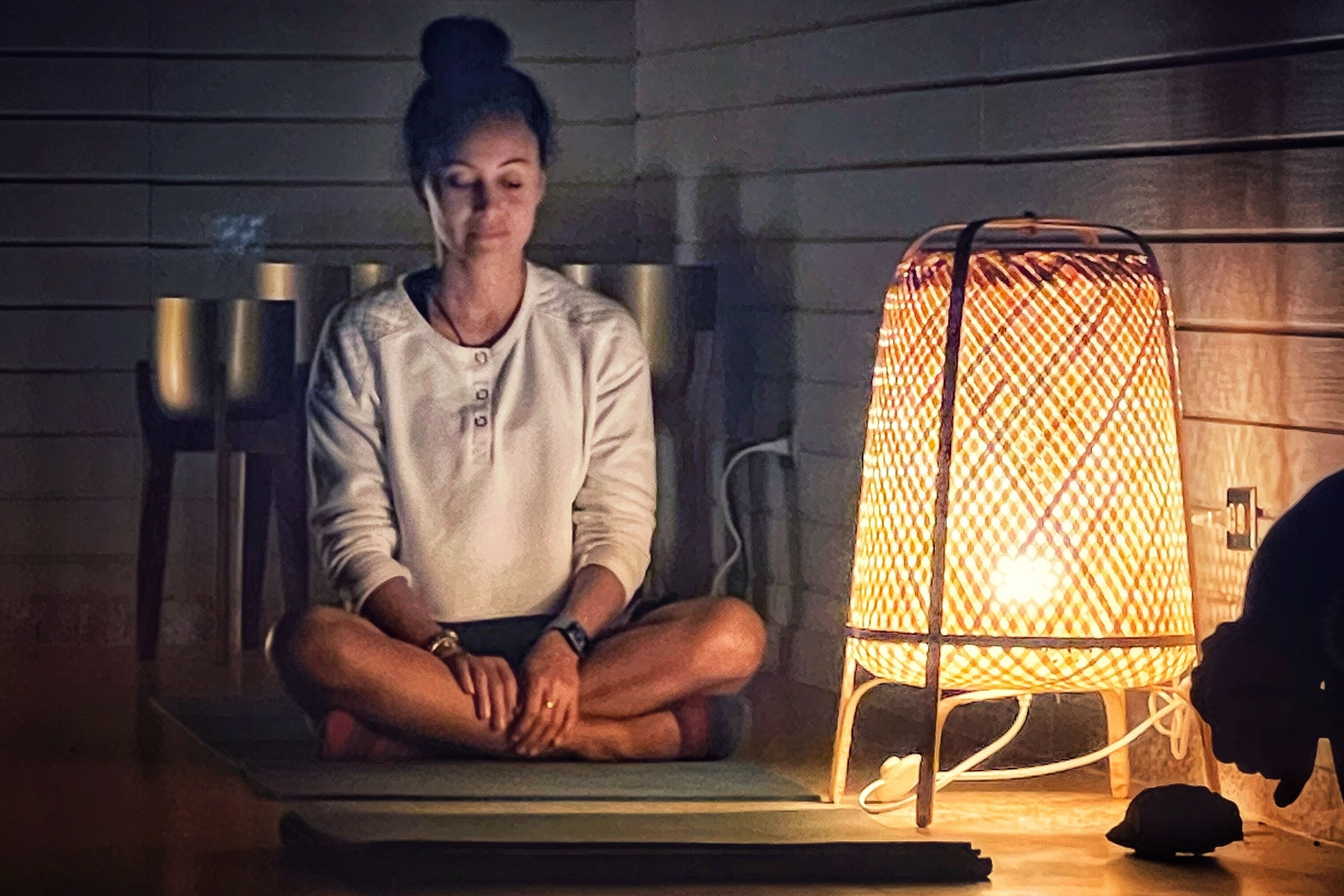 A person sitting on a mat in a dimly lit room with a patterned lamp.