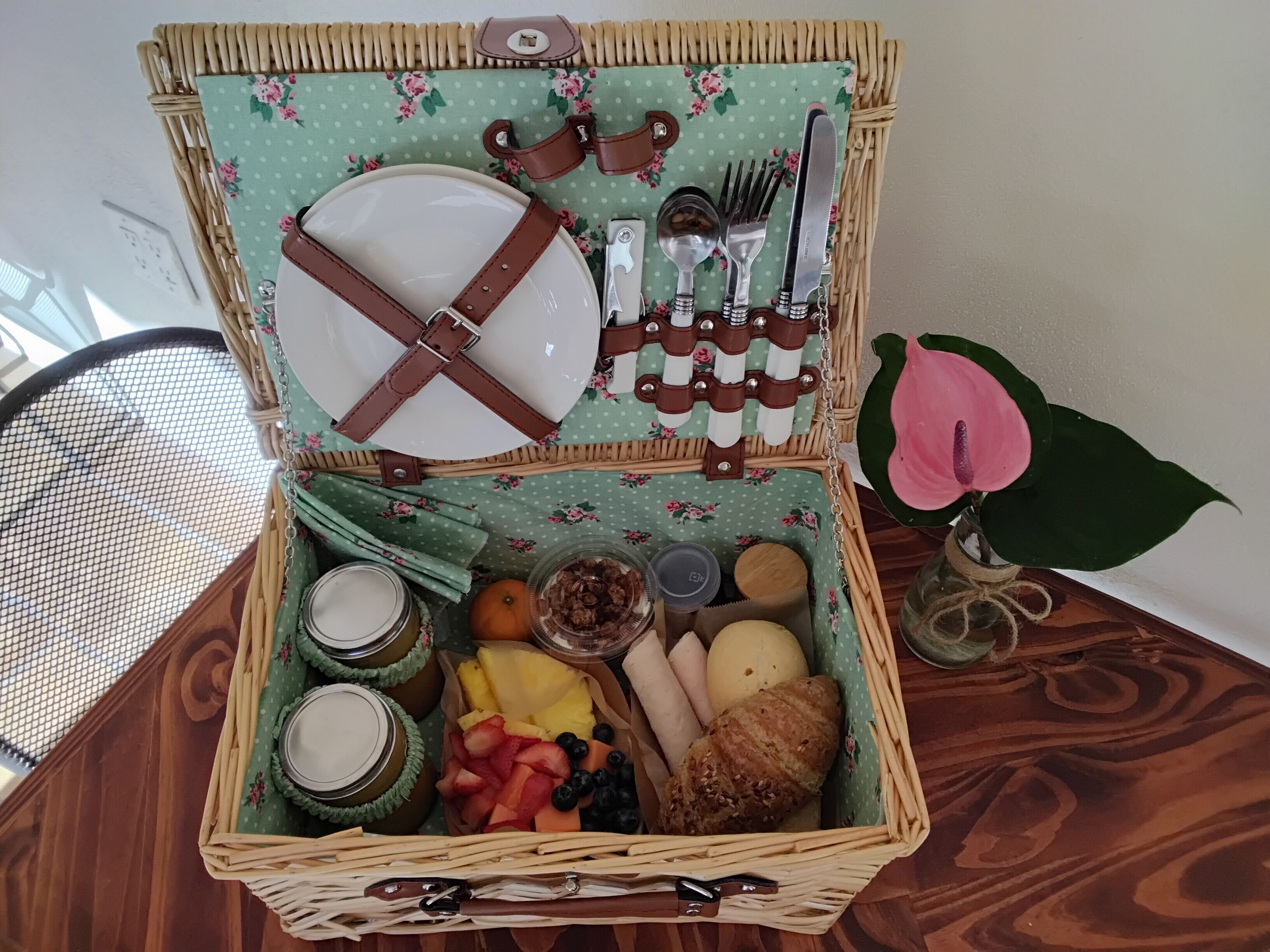 A wicker picnic basket with a floral tablecloth, containing a plate, cutlery, jars, fruits, and a bread roll.