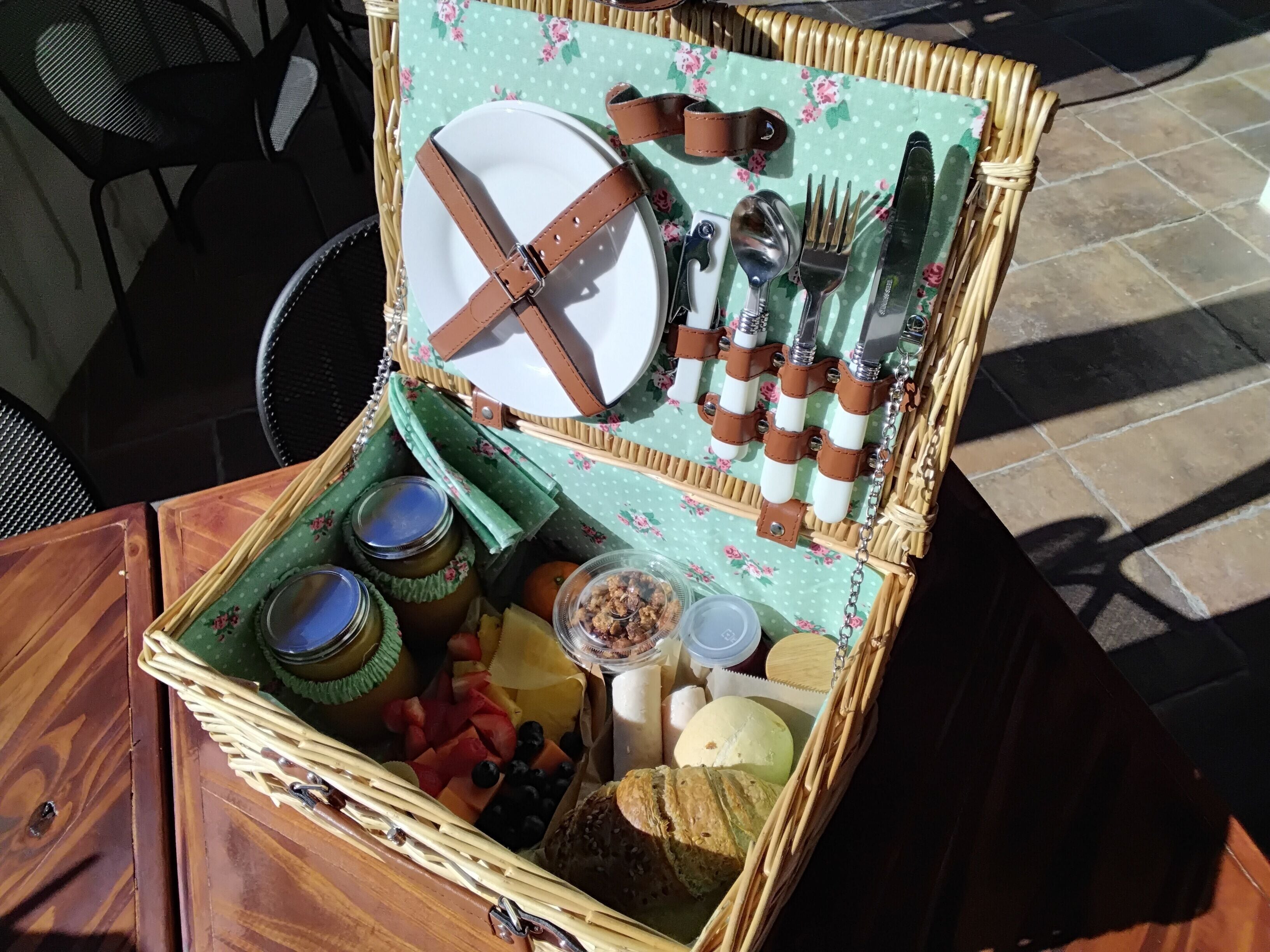A wicker picnic basket with a floral tablecloth, containing a plate, cutlery, jars, fruits, and bread.