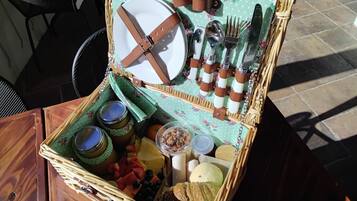 A wicker picnic basket with a floral tablecloth, containing a plate, cutlery, jars, fruits, and bread.