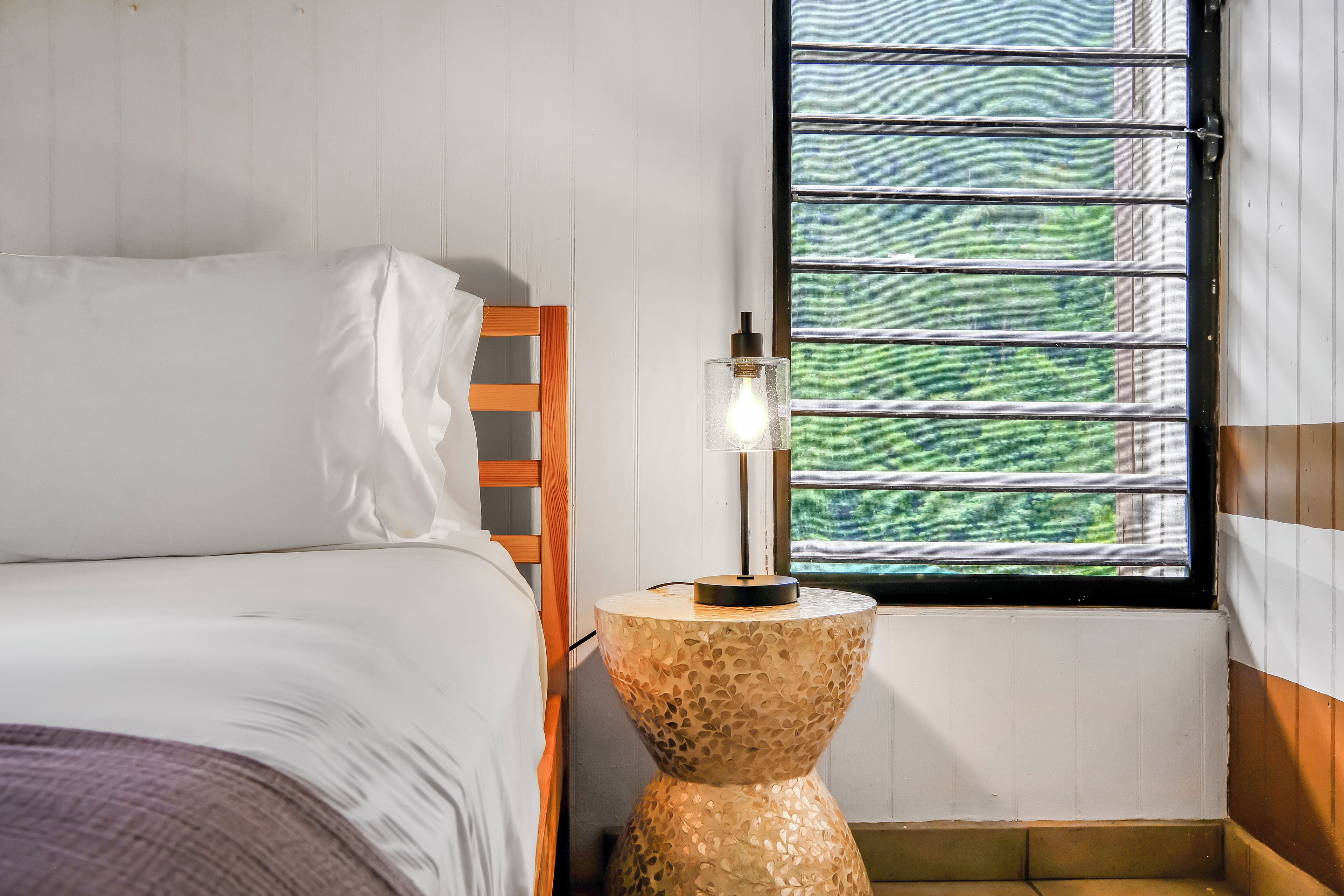 A bedroom with a bed, a wooden headboard, a round side table with a light bulb, and a window with a view of greenery.