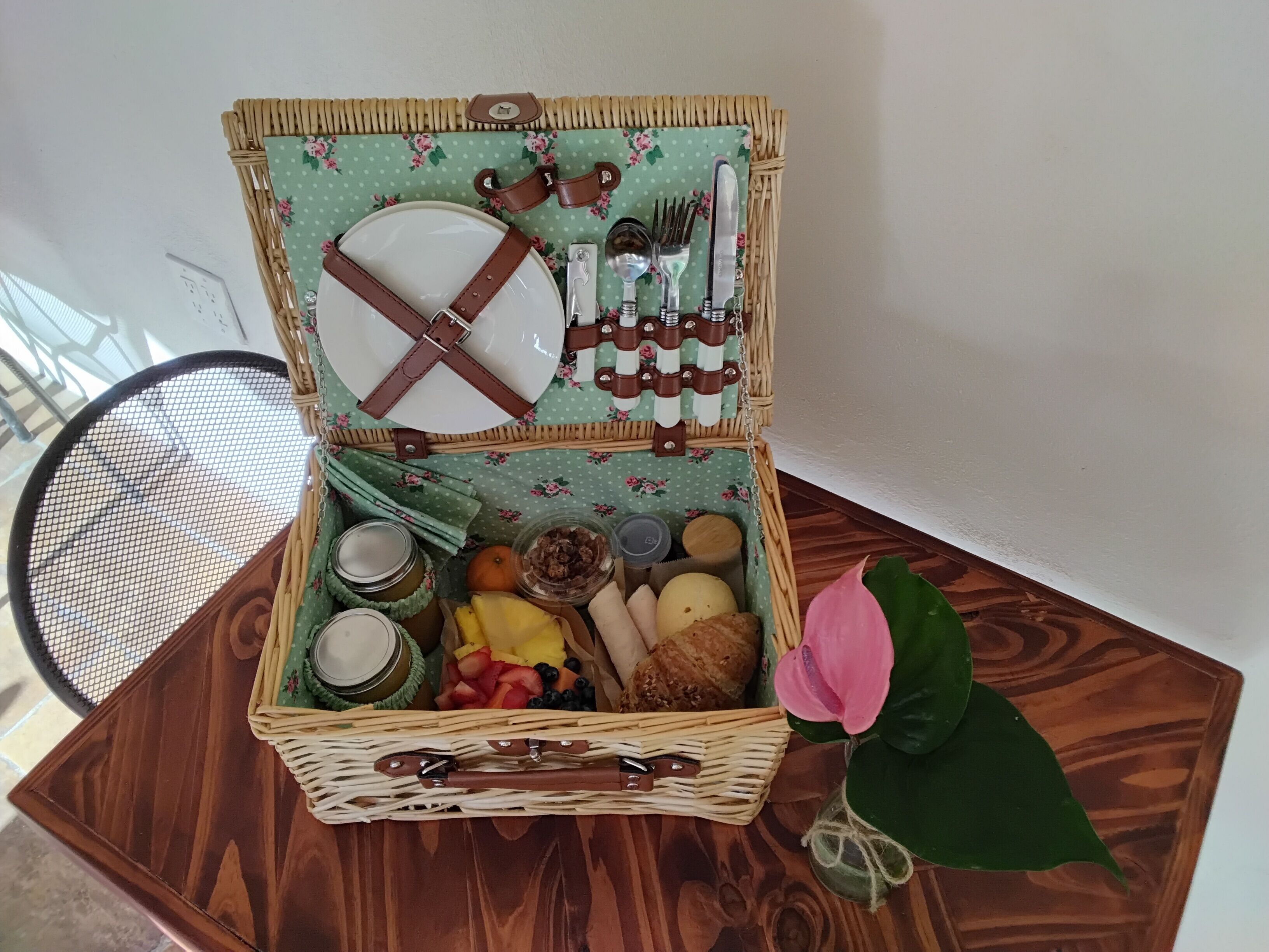 An open wicker picnic basket with a floral pattern, containing a plate, cutlery, jars, a bowl of fruit, and a pink flower.