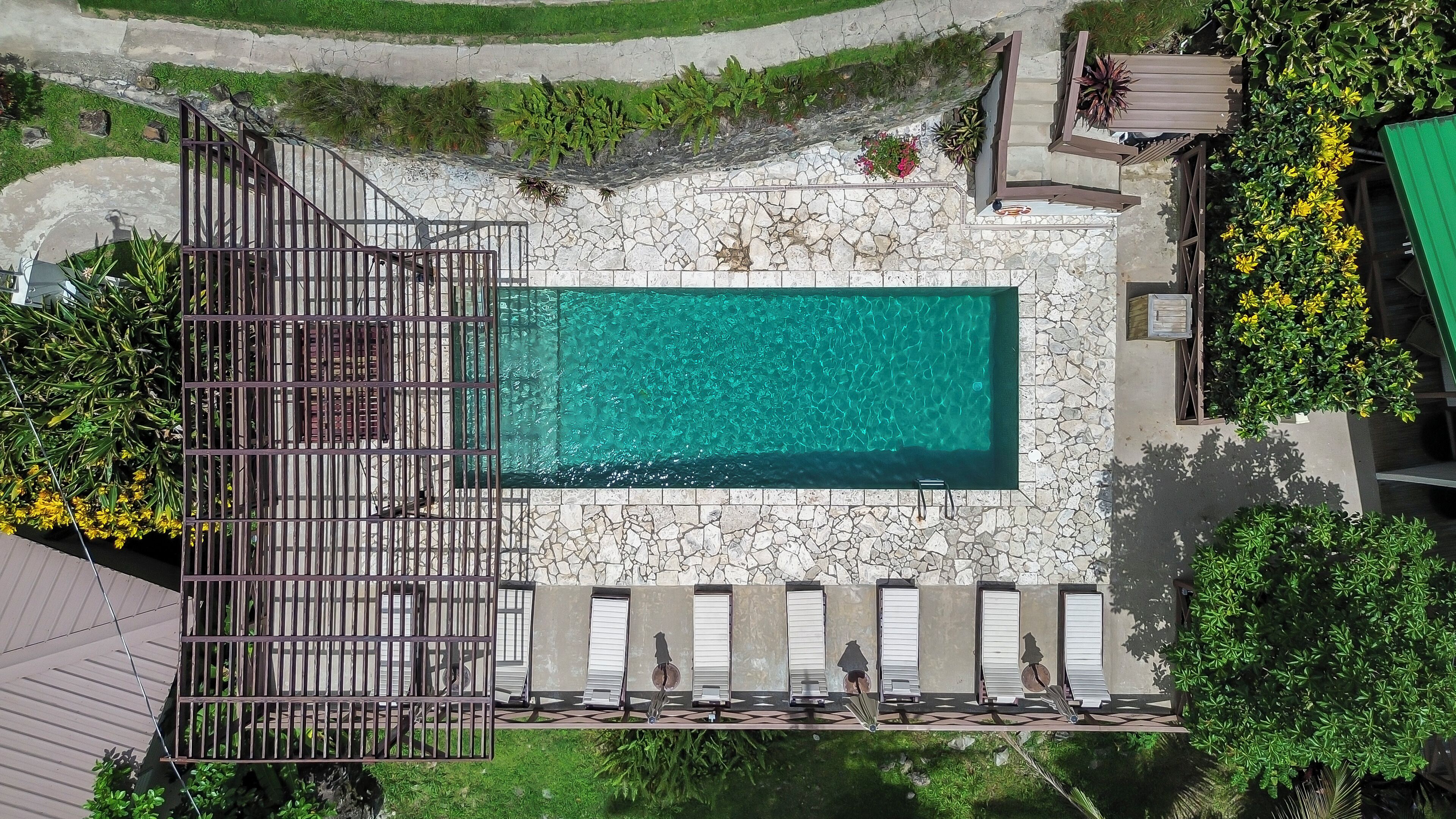 An aerial view of a swimming pool with a stone deck, surrounded by greenery and a wooden fence.