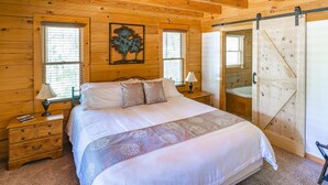 A wooden cabin bedroom with a bed, two nightstands, a lamp, a painting, and a sliding door to a bathroom. - Hillside Haven | Luxe Hot Tub | Cornhole | Jenga (Whitwell)