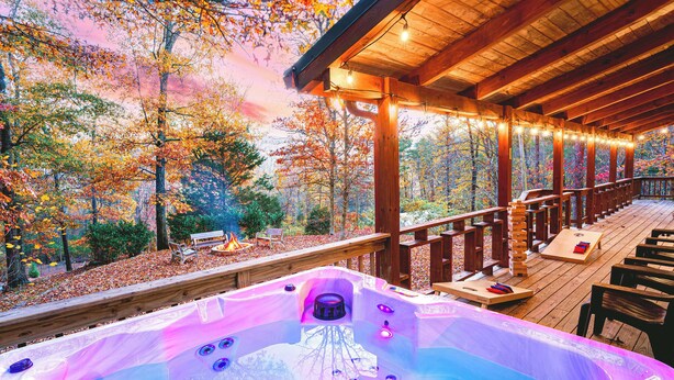 A hot tub with a wooden deck, surrounded by autumn foliage and a wooden pergola with string lights. - Hillside Haven | Luxe Hot Tub | Cornhole | Jenga (Whitwell)