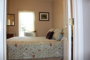 Ein Schlafzimmer mit blumigem Bettbezug, einem Fenster mit Vorhängen, einem gerahmten Bild an der Wand und einem Nachttisch.