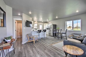 A modern living room with a grey sofa, a wooden coffee table, and a kitchen area with a fridge and bar stools. - Bright + Modern Augusta Home - Walk to Town! (Augusta)