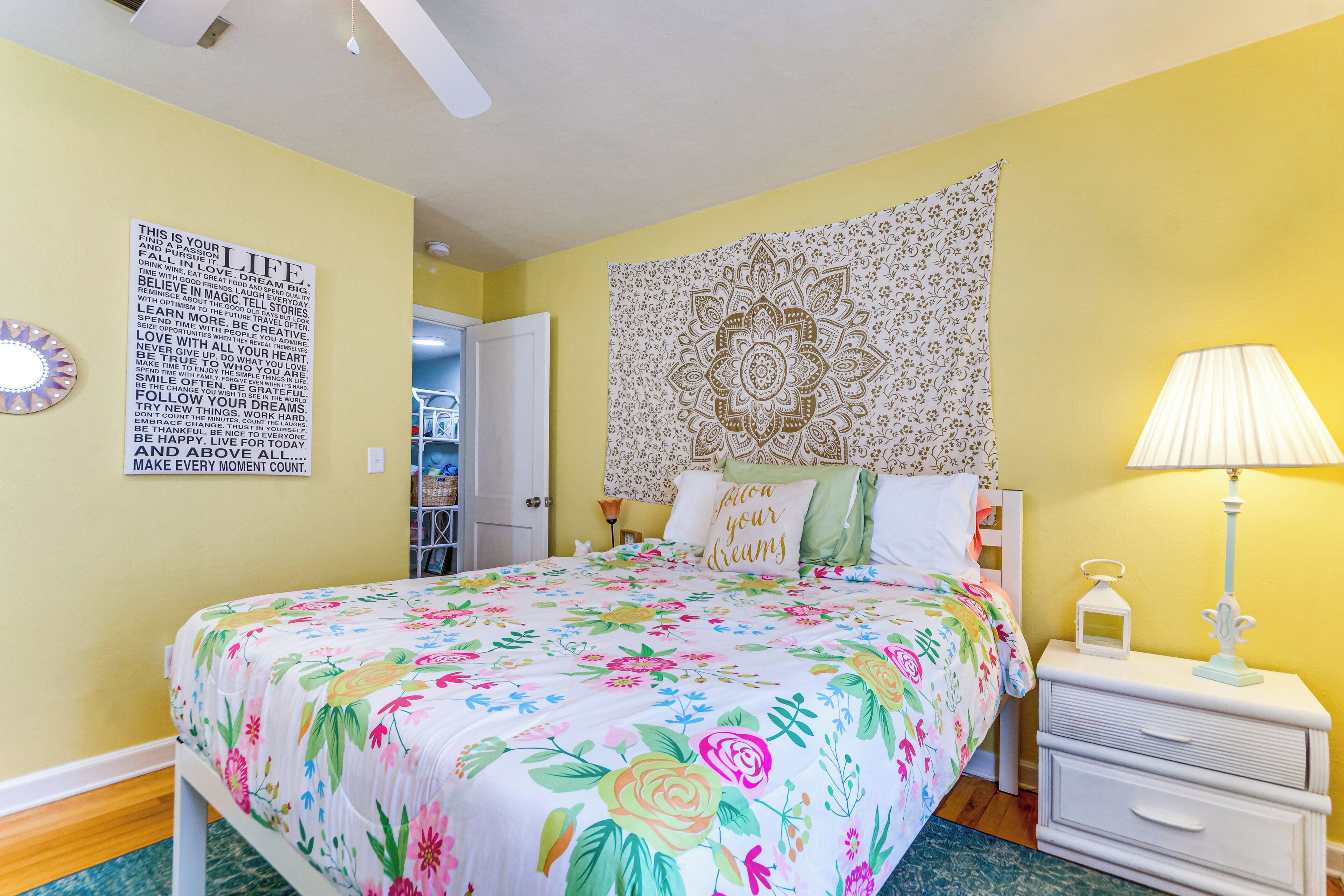 Une chambre à coucher avec une couvre-lit à motifs floraux, une tête de lit en tapisserie, une lampe de chevet et une citation inspirante sur le mur.